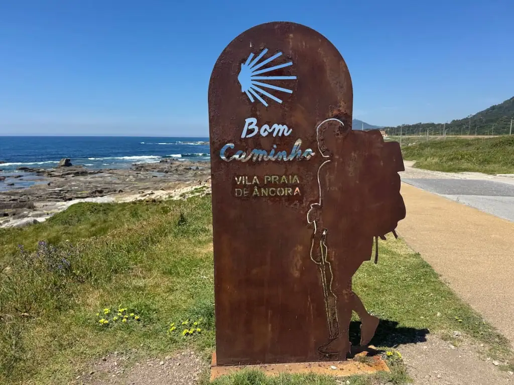 Brown metal sign with the Camino scallop shell symbol and a pilgrim that says "Bom Caminho Vila Praia de Âncora" nex to a path along the coast.