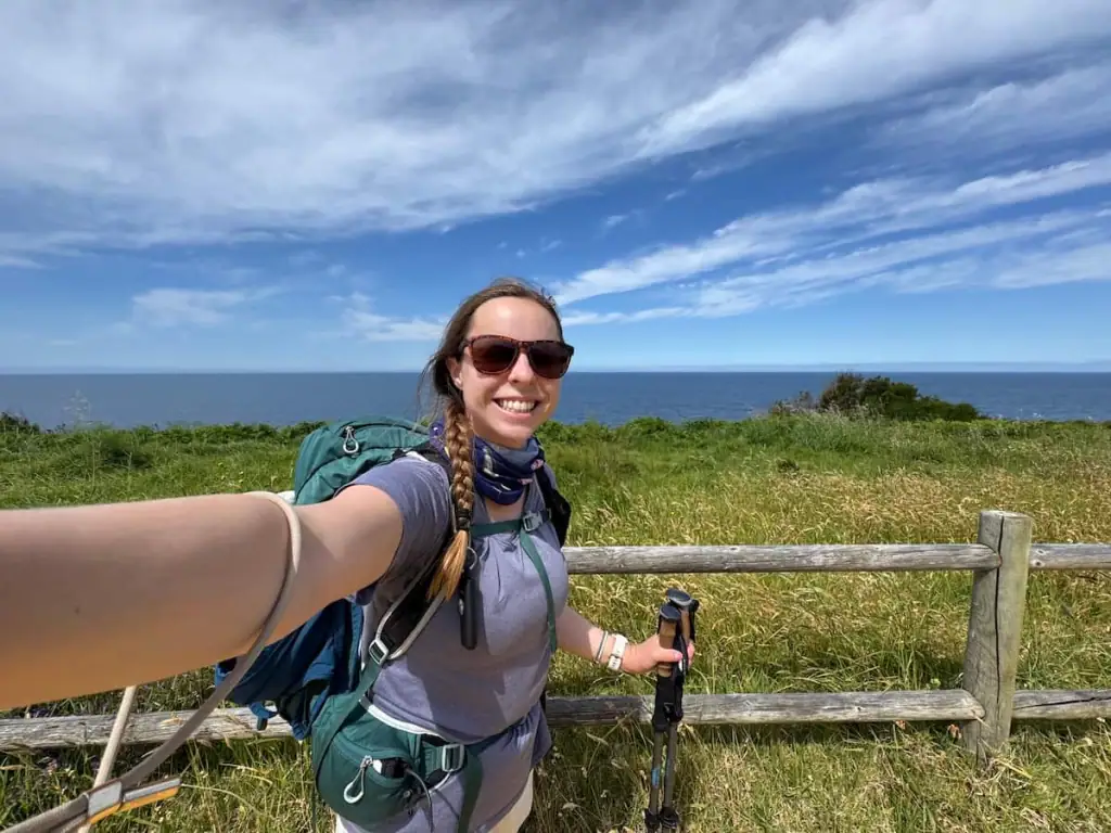 Mikala smiling in a selfie while hiking on the Camino Portugués Coastal route with a wooden fence and grass behind her with the ocean behind.