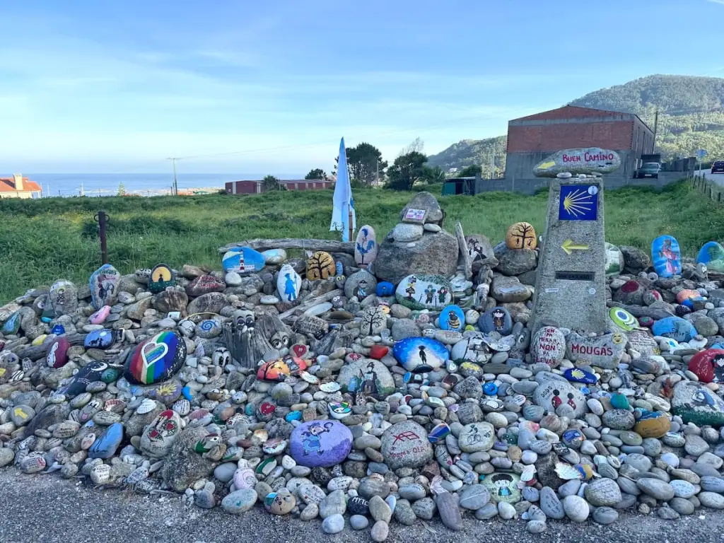 Pile of painted rocks next to a Camino trail marker with a building and the ocean in the distance north of Viladesuso on the Camino Portugués Coastal route.