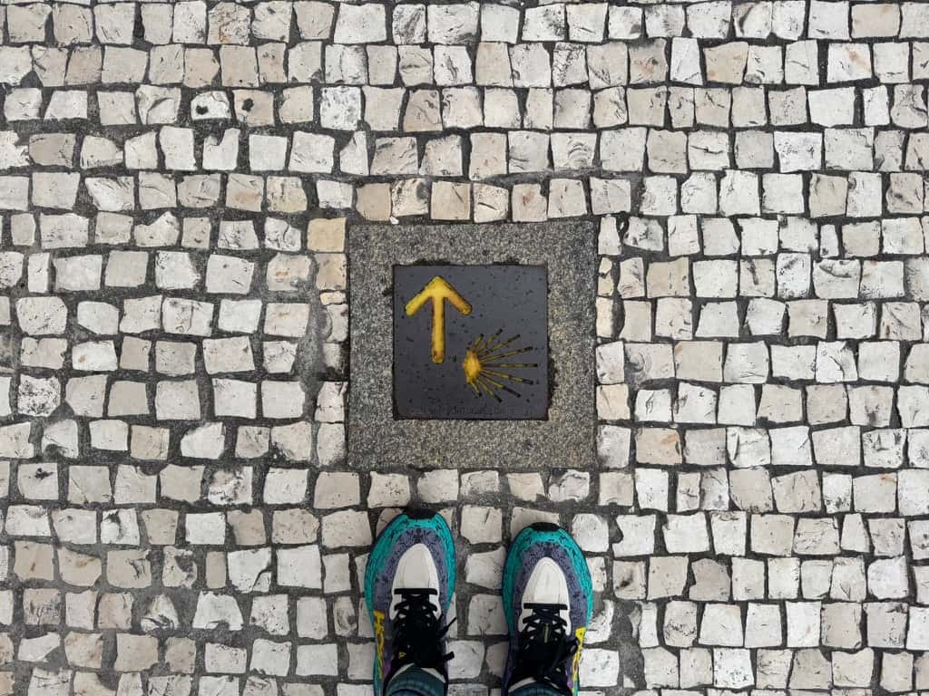 Mikala's feet standing on a cobblestone path in front of a Camino trail marker pointing the way on the Camino Portugués.