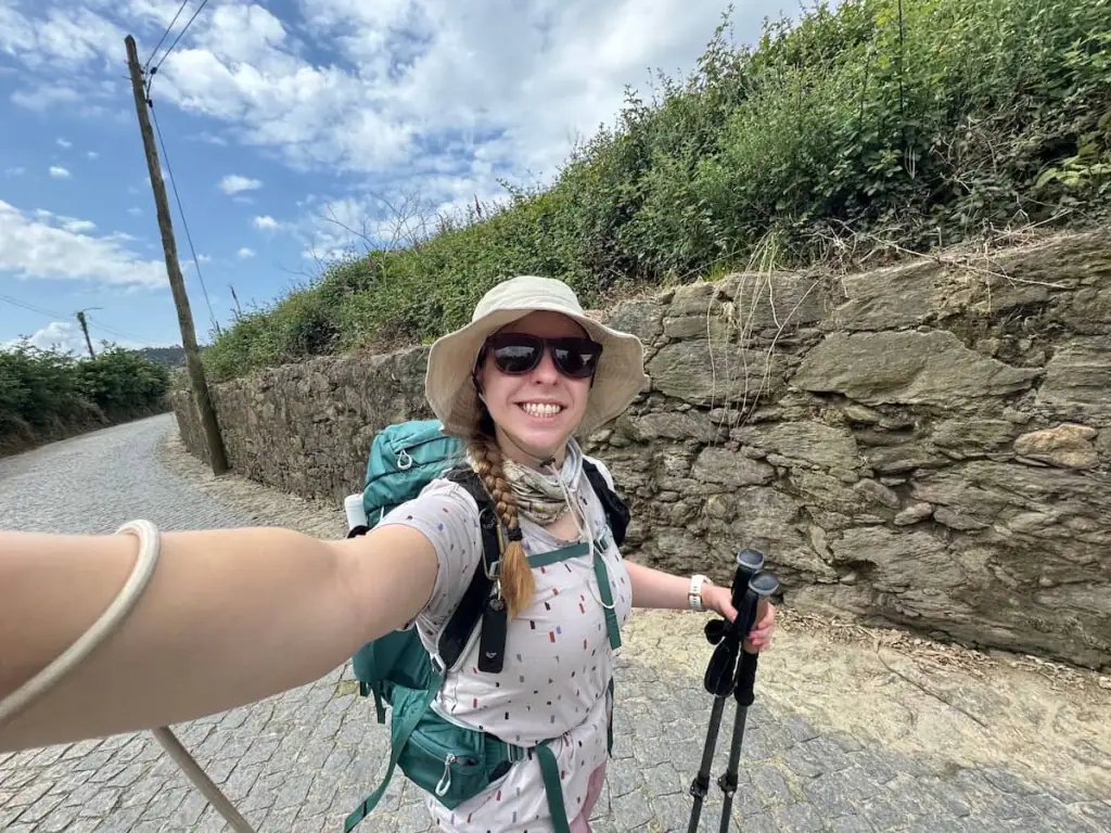 Mikala smiling in a selfie walking along the road in Mosteiró with a stone wall and bushes behind her on the Camino Portugués Central route.