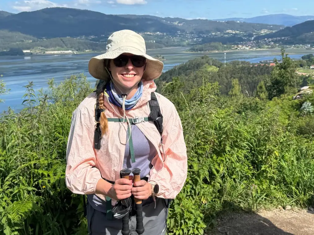 Mikala wearing a jacket and hat and holding her hiking poles while smiling with a river and mountains in the background.