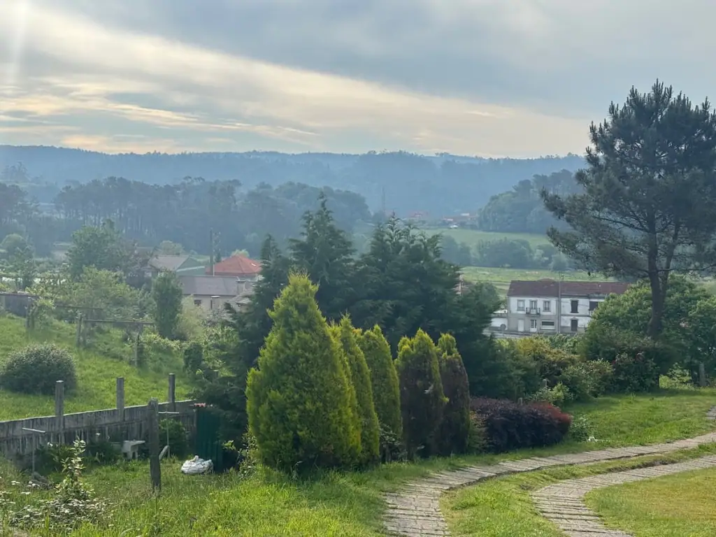 View of tall green trees, a few houses, and mountains in the distance on a semi-cloudy morning in Santa Mariña de Carracedo on the Camino Portugués Central route.