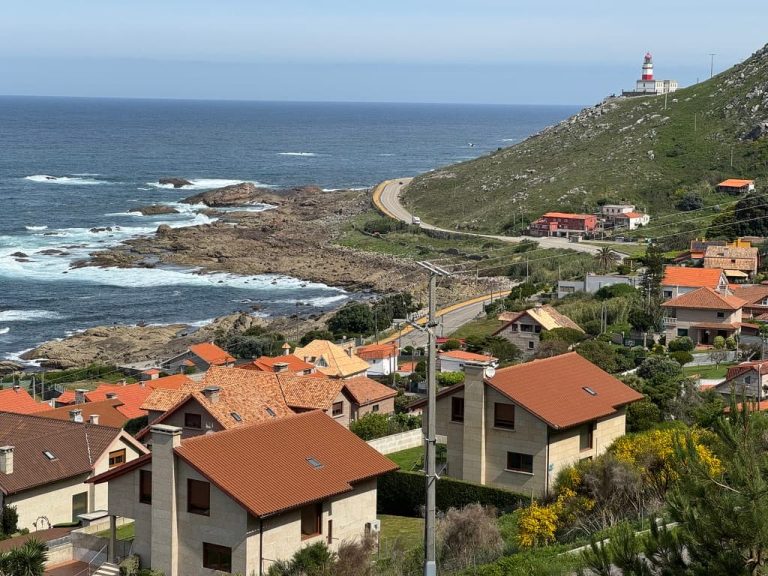 Small village and road along the coastline in Oia, Spain outside of Baiona with a small red and white lighthouse on a hill in the distance.