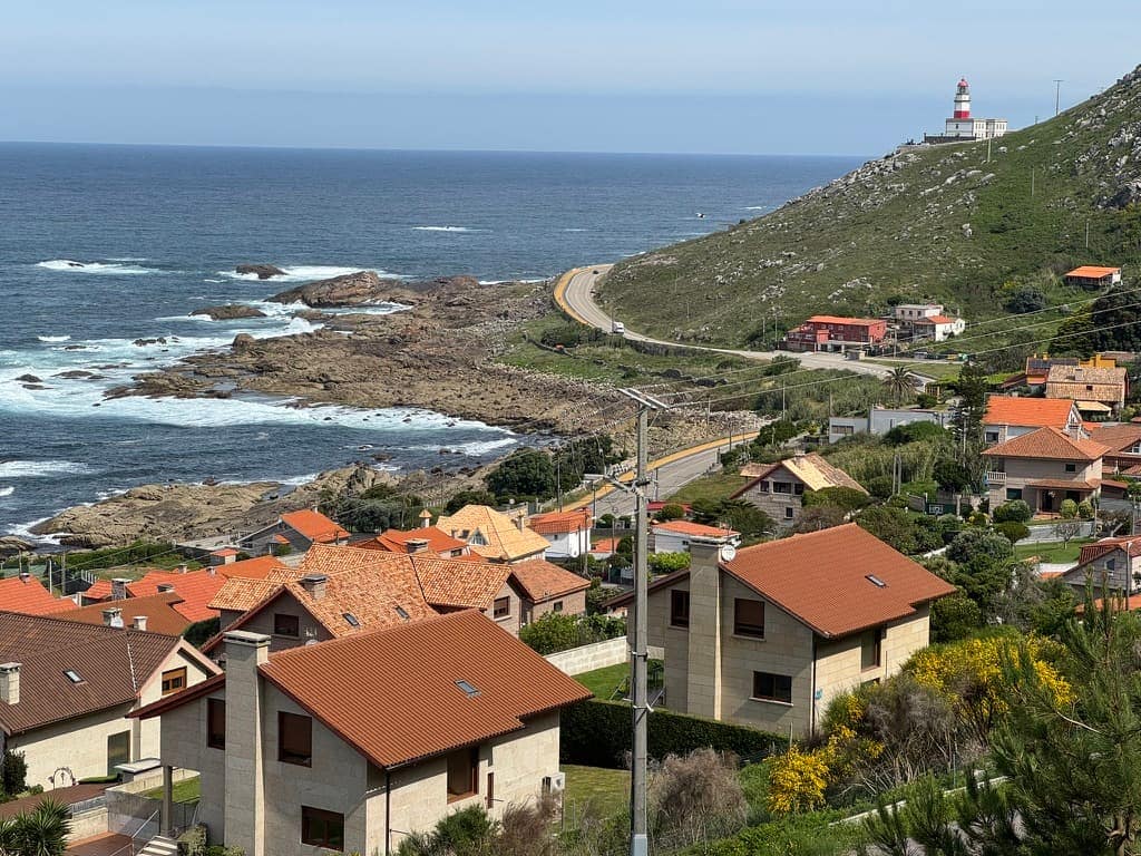 Small village and road along the coastline in Oia, Spain outside of Baiona with a small red and white lighthouse on a hill in the distance.