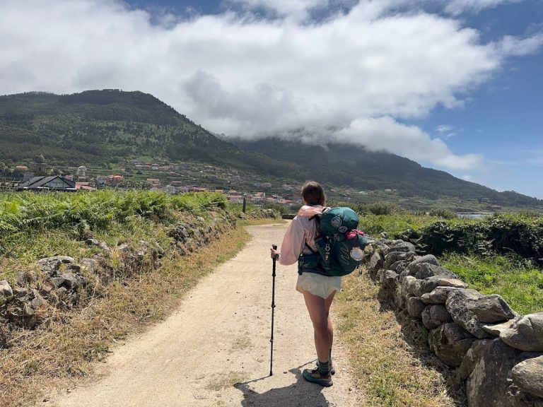 Mikala standing on a dirt path with her backpack and hiking poles looking away from the camera back towards a mountain and town in the distance while hiking solo on the Camino Portugués Coastal route.