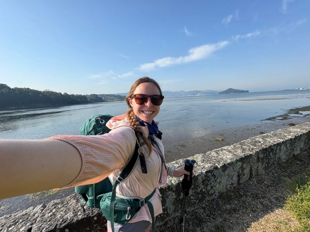 Mikala taking a selfie smiling with water and small mountains behind her while on the way to Combarro on the Camino Portugués Spiritual Variant.
