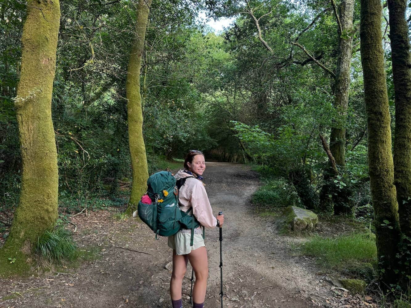 Mikala walking in a lush green forest along the Spiritual Variant on the Camino Portugués and turning back to smile at the camera.