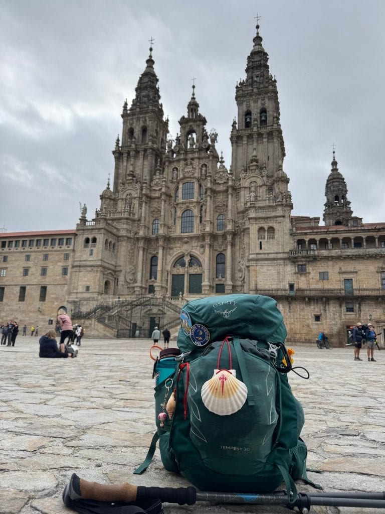 Mikala's green Camino backpack with shell and hiking poles sitting in the cathedral square in Santiago de Compostela with the cathedral in the background.