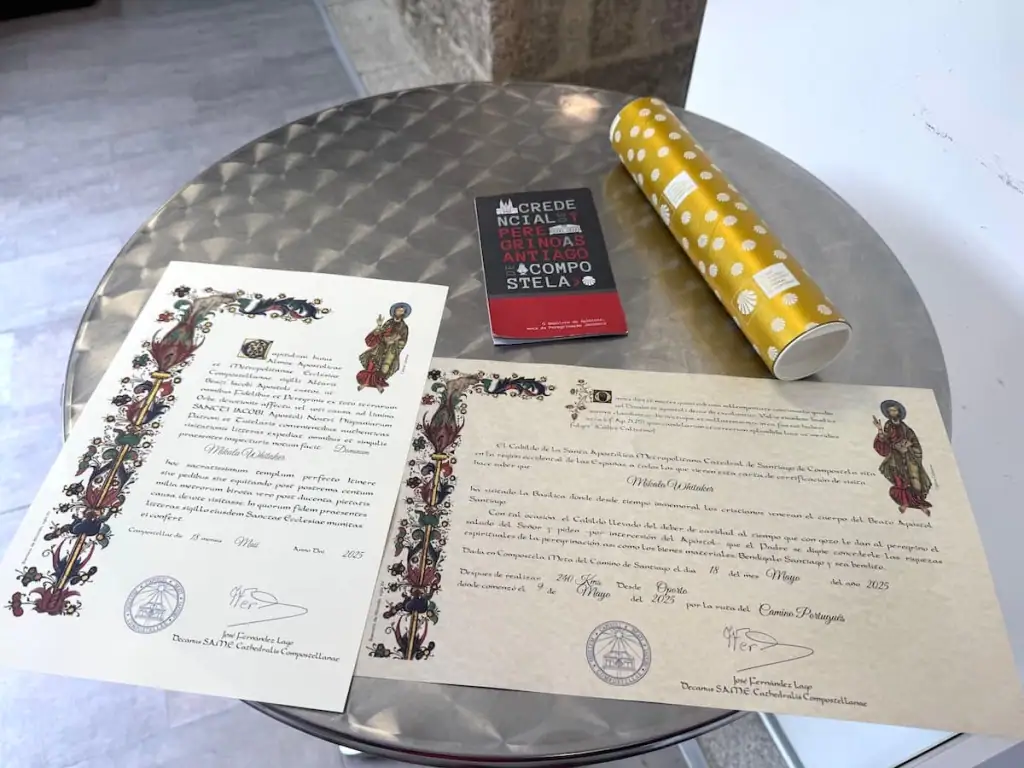 Camino de Santiago Compostela certificate, distance certificate, pilgrim's passport, and a tube to hold them on a silver table.