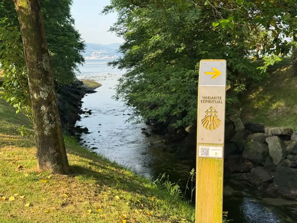Silver trail marker for the Camino Spiritual Variant with trees, water, and a small mountain in the background.