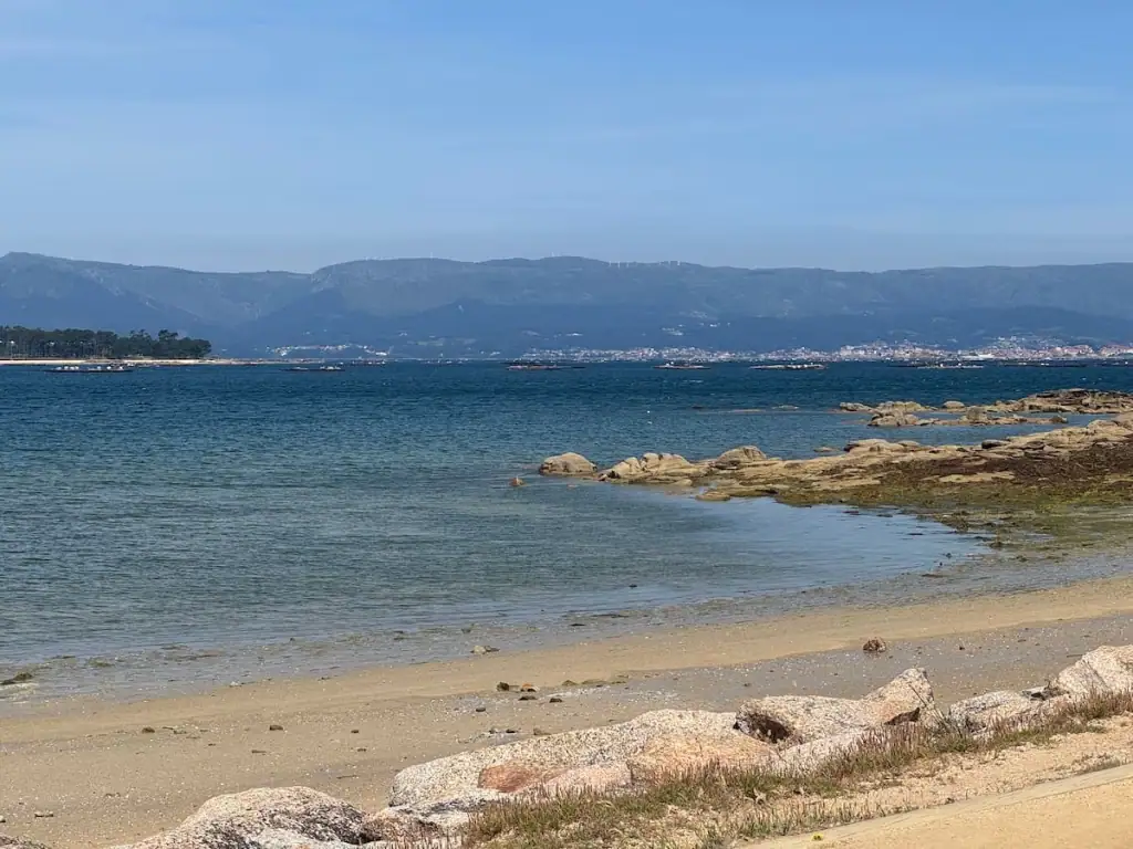 Sandy beach with some rocky areas sticking out into vibrant dark blue water with mountains in the distance. This is along the Spiritual Variant shortly before reaching Vilanova de Arousa.