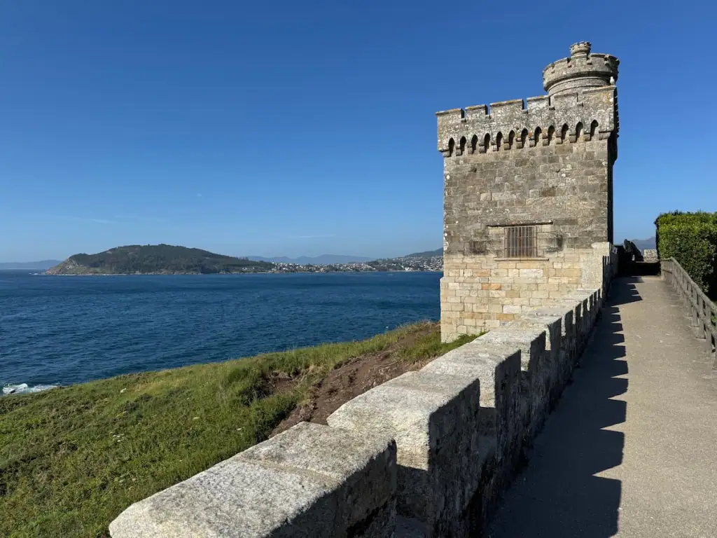 Fortress wall and tower in Baiona, Spain with green grass and the ocean below and another green peninsula visible in the distance on a sunny day with blue skies.