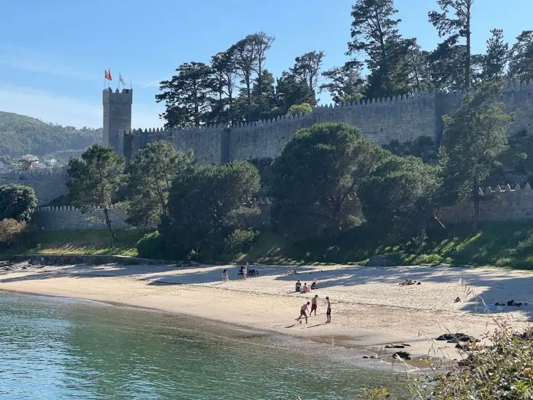 Praia da Barbeira beach in Baiona, Spain along the Camino de Santiago with the fortress and trees visible behind the beach, blue green water, and clear blue skies.