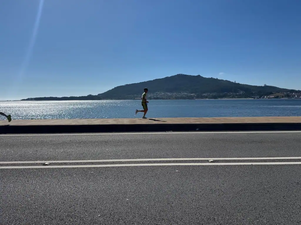 Runner on a paved path in Caminha between the water and a road with a mountain in the distance.