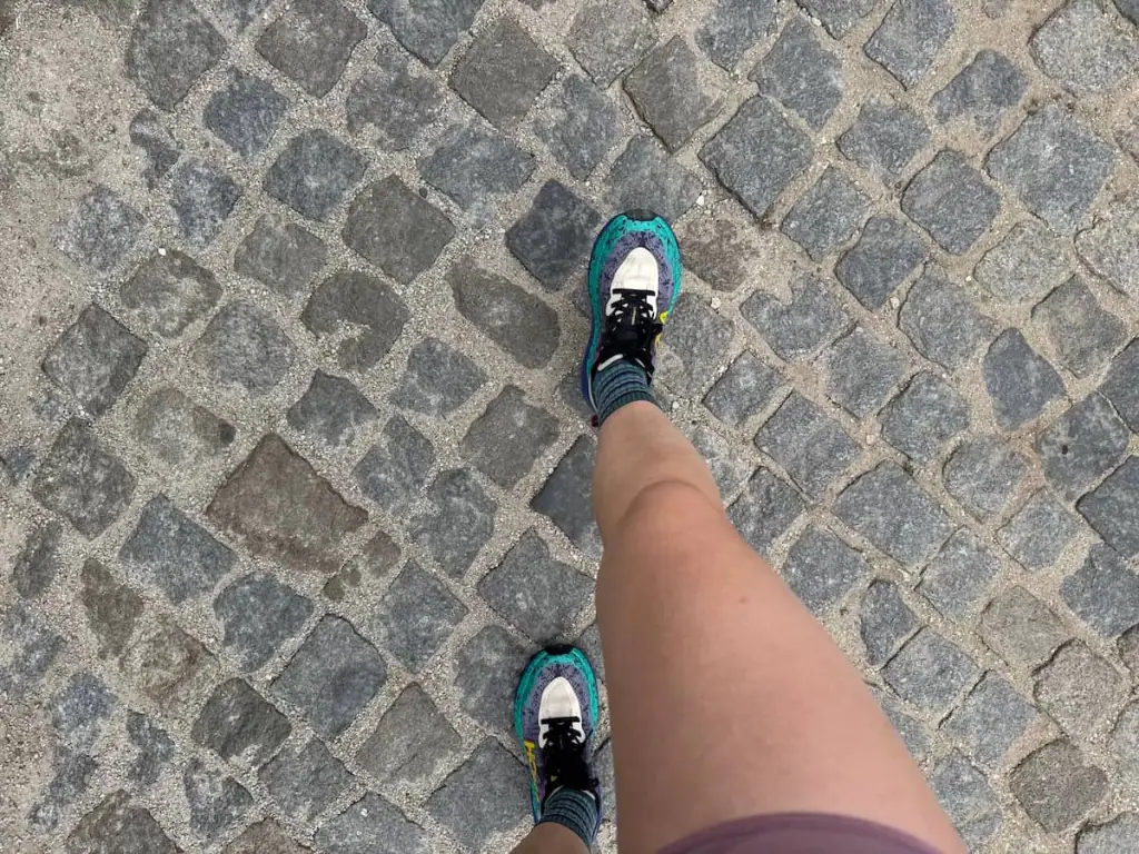 View from above of two legs walking along a cobblestone road on the Camino Portugués with Hoka Speegoat shoes on the feet.