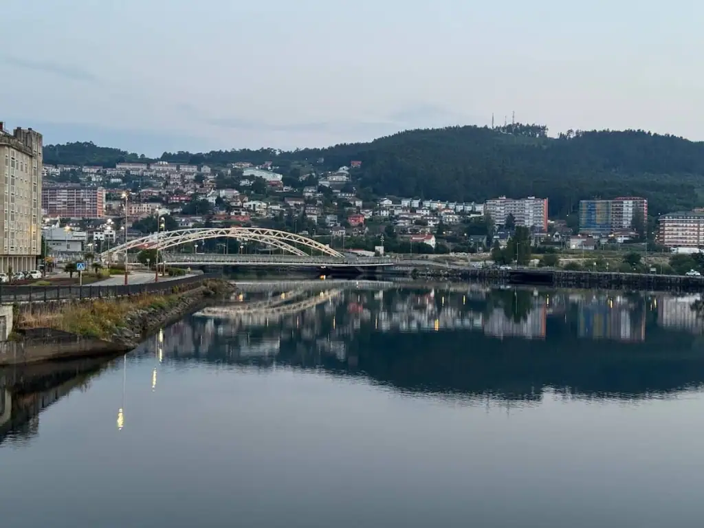 View of Pontevedra on the Camino de Santiago from the Burgo Bridge over the river with another bridge visible, colorful buildings on the right, and a mountain in the distance at sunrise.