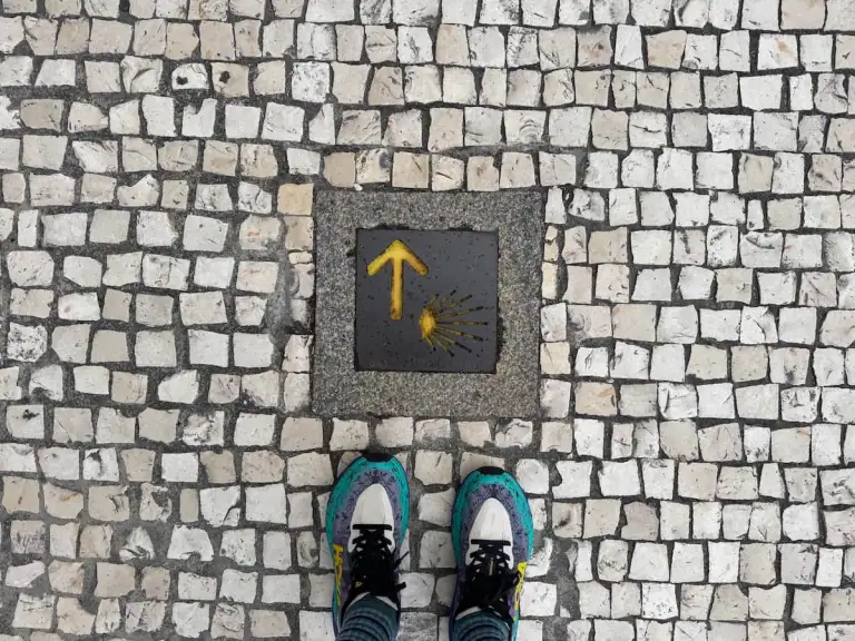Mikala's feet standing on a cobblestone path in front of a Camino trail marker pointing the way on the Camino Portugués.