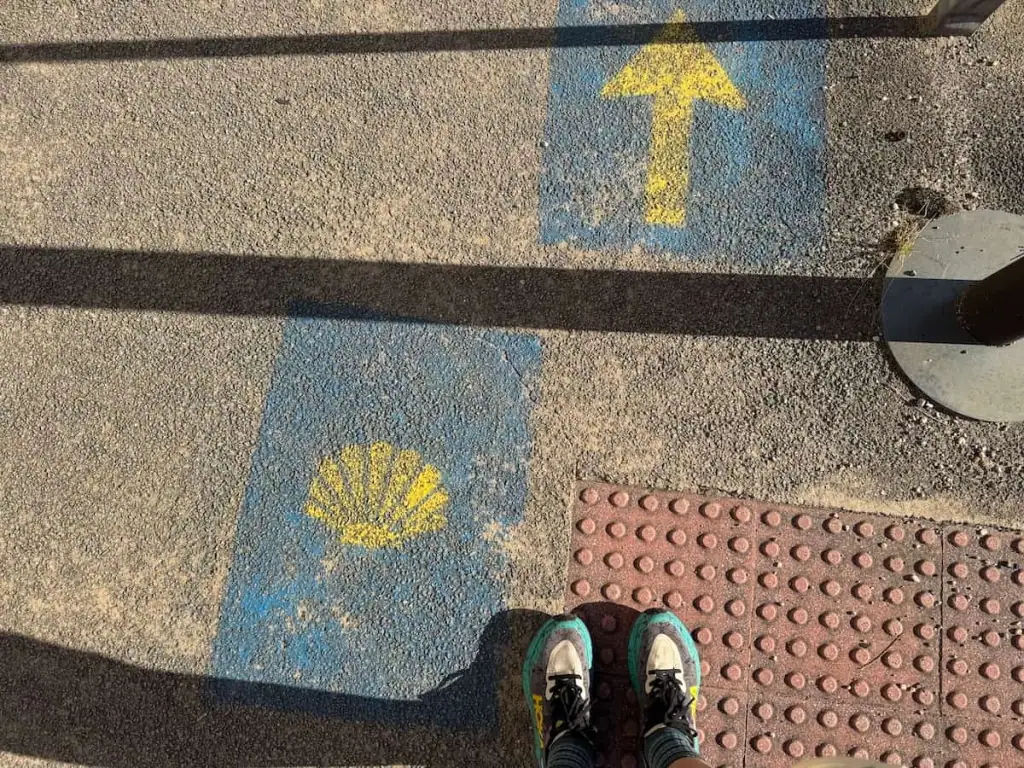 Two feet wearing Hoka Speedgoats visible on pavement that has a yellow Camino scallop shell and a yellow arrow printed on it to point the way.