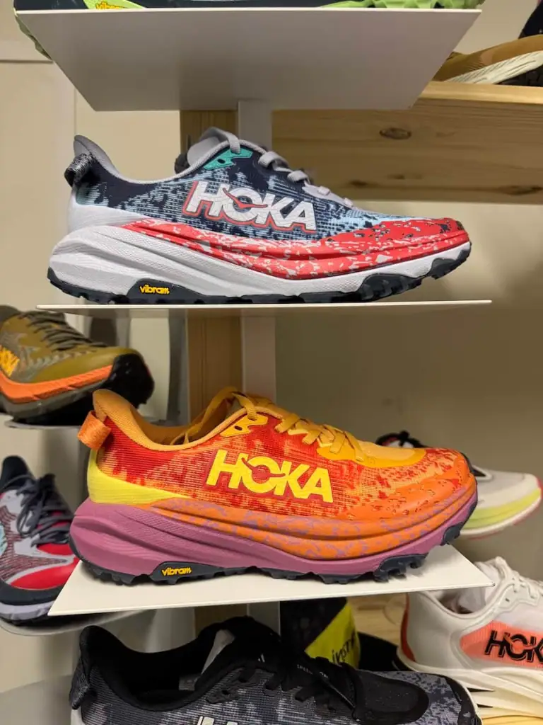 Two Hoka Speedgoat shoes on shelves with one above the other. The bottom is a bright orange color with pink and yellow accents while the top is blue with red and white accents.