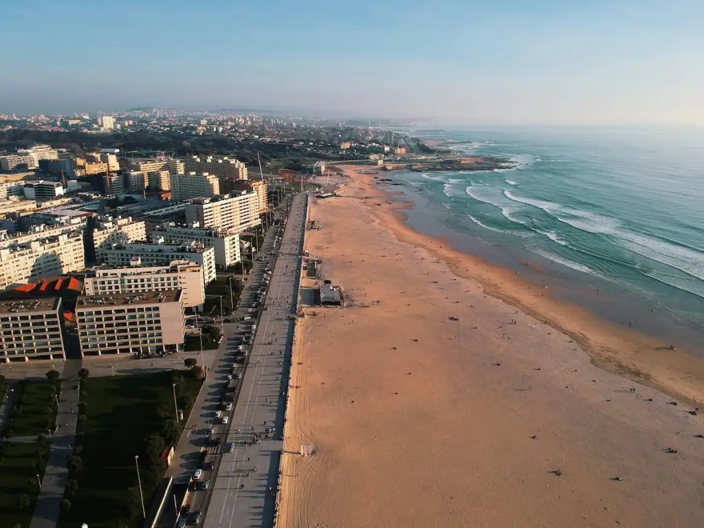 Overhead view shortly before sunset of Matosinhos, Portugal with the light blue ocean on the left, sand in the middle, and road and buildings on the left.