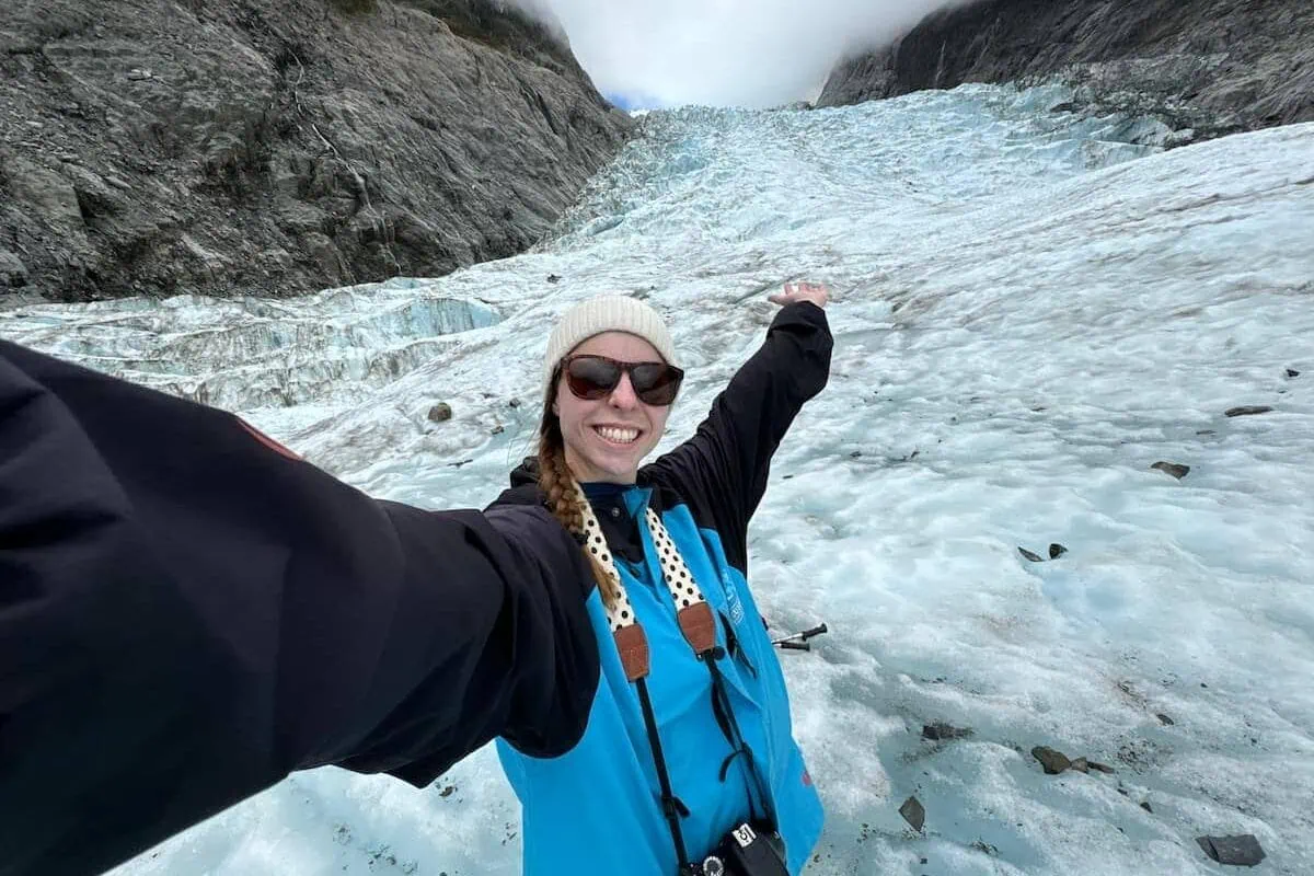 Mikala taking a selfie with her hand up in the air in front of Fox Glacier while on a heli hiking tour in New Zealand.