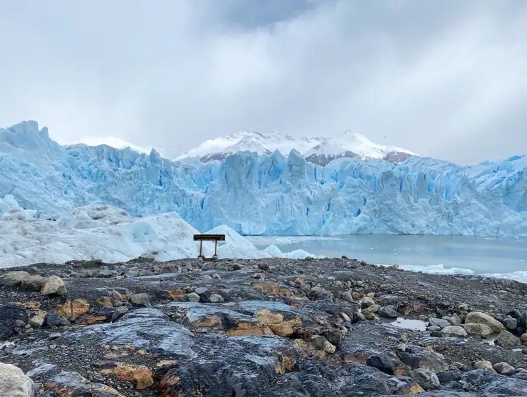 View of the Perito Moreno Glacier in Patagonia, Argentina, with icy blue walls rising behind a rocky foreground and a small wooden sign overlooking the glacier. Overcast skies and snow-covered mountains frame the dramatic landscape, capturing a must-see stop on any Argentina itinerary.
