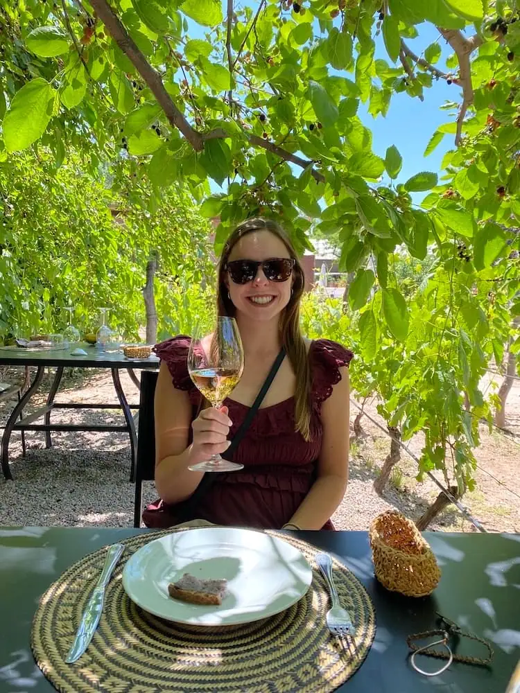 Woman sitting at a table surrounded by trees and grapevines at a vineyard in the Mendoza, Argentina area smiling holding up a glass of red wine.