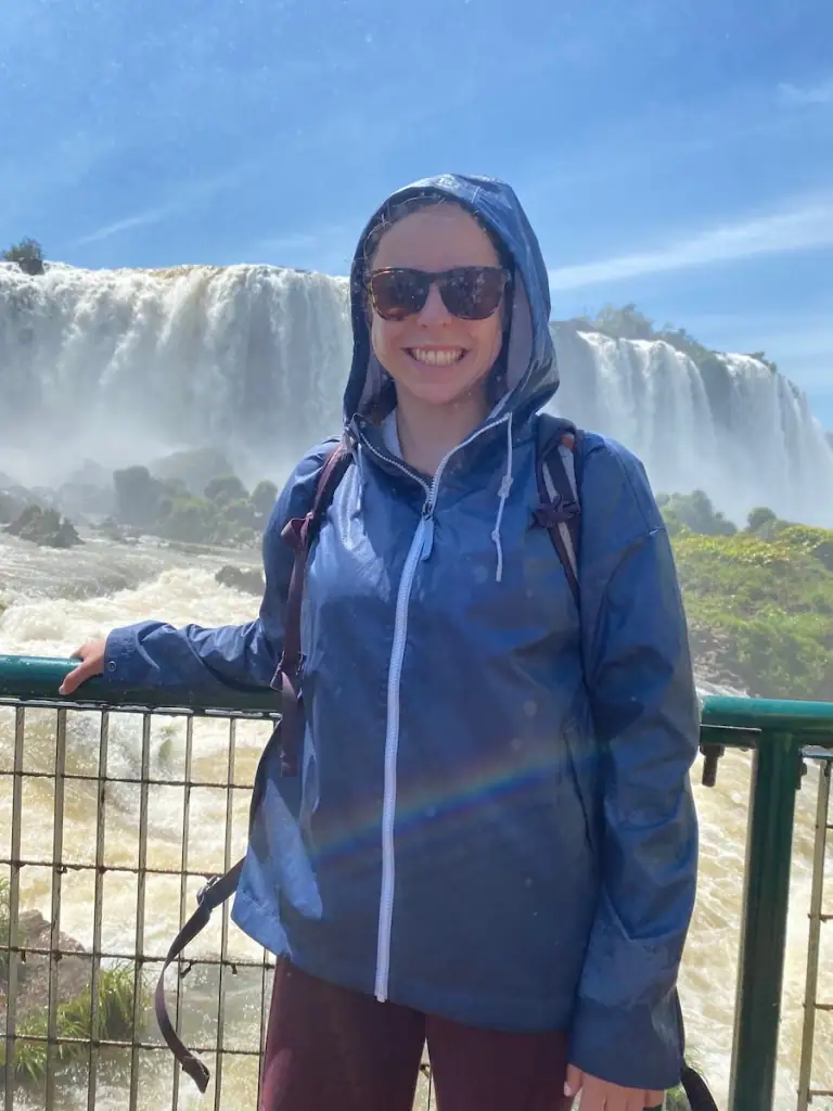 Woman in a rain jacket with the hood up smiling while on a walkway right in front of Iguazú Falls on the Brazilian side.