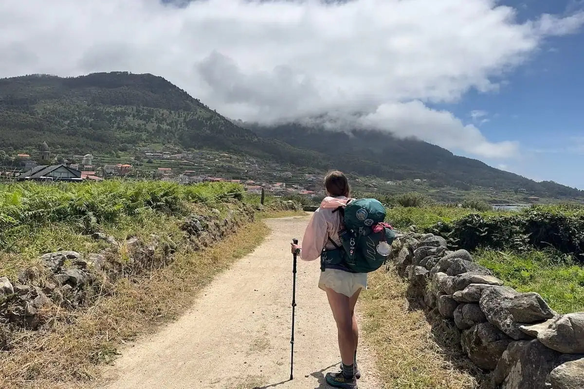 Mikala standing on a dirt path with her backpack and hiking poles looking away from the camera back towards a mountain and town in the distance while hiking solo on the Camino Portugués Coastal route.