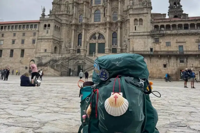 Mikala's green Camino backpack with shell and hiking poles sitting in the cathedral square in Santiago de Compostela with the cathedral in the background.
