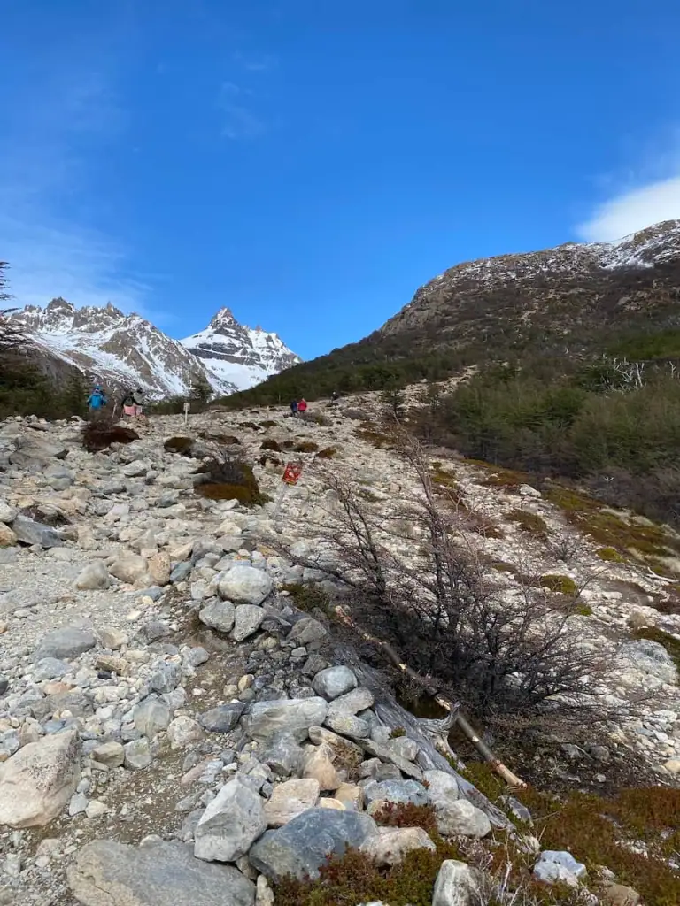 Steep rocky mountain trail on the Mount Fitz Roy hike ascending toward snowy peaks with hikers in the distance under a bright blue sky.