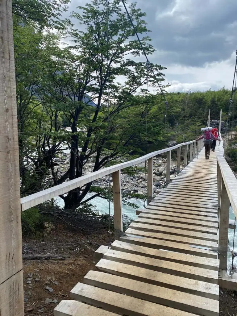 Two hikers cross a wooden suspension bridge over a rushing river, surrounded by forest and mountain scenery along the French Valley hike in Torres del Paine.