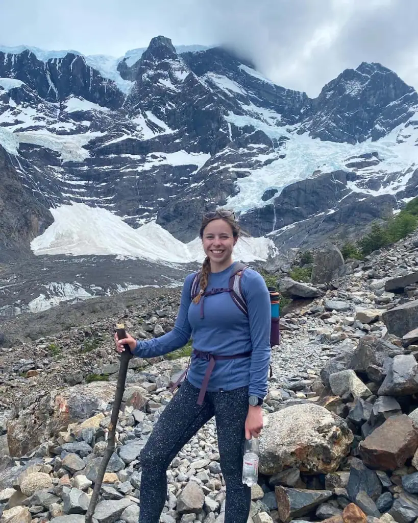 Mikala smiling with a hiking stick at the Mirador Francés on the French Valley hike in Torres del Paine with a glacier and jagged snowy peaks rising behind her.