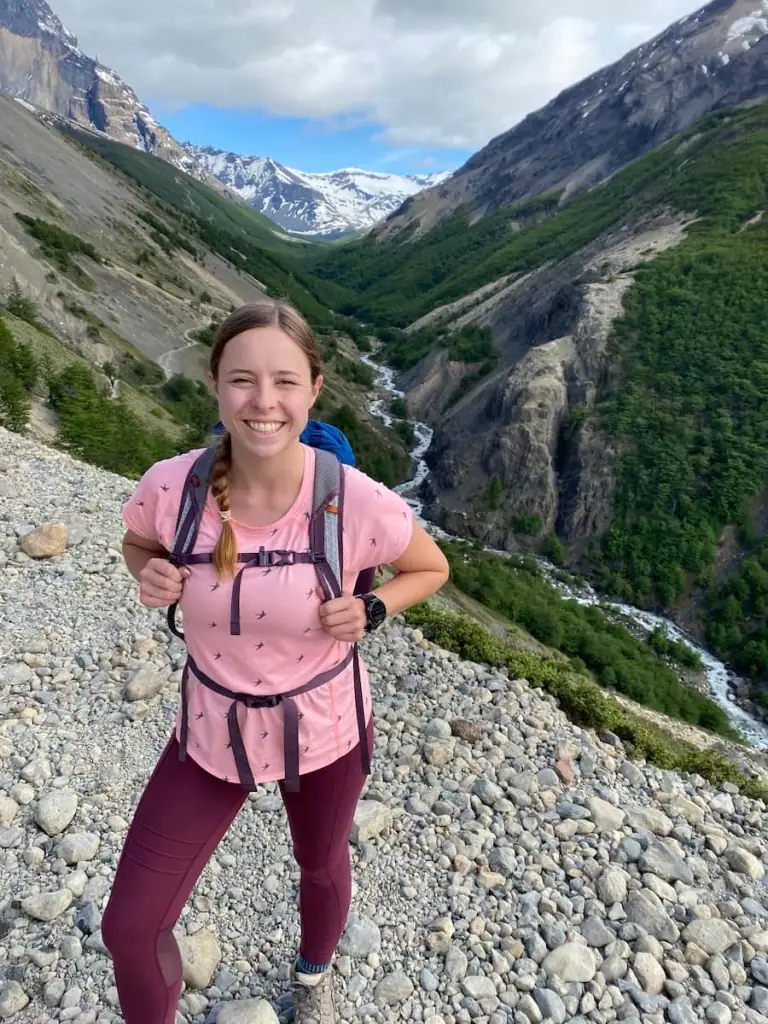 Mikala smiling in a pink shirt and maroon leggings while hiking on a rocky trail section of the Mirador las Torres hike in Torres del Paine with snow-capped mountains and a winding river in the background.
