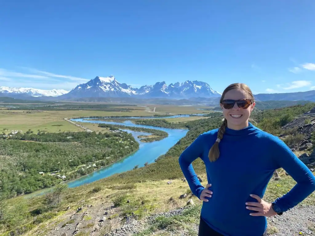 Mikala smiling in a blue sweater above a winding turquoise river with the sharp peaks of Torres del Paine rising in the background.