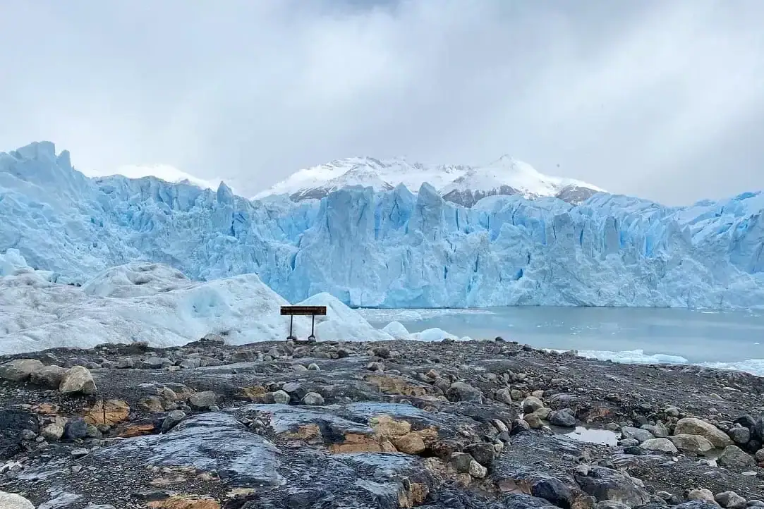View of the Perito Moreno Glacier in Patagonia, Argentina, with icy blue walls rising behind a rocky foreground and a small wooden sign overlooking the glacier. Overcast skies and snow-covered mountains frame the dramatic landscape, capturing a must-see stop on any Argentina itinerary.