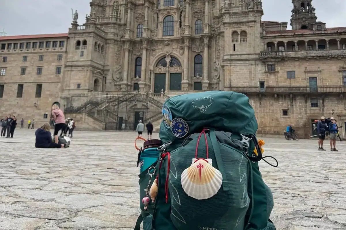 Mikala's green Camino backpack with shell and hiking poles sitting in the cathedral square in Santiago de Compostela with the cathedral in the background.