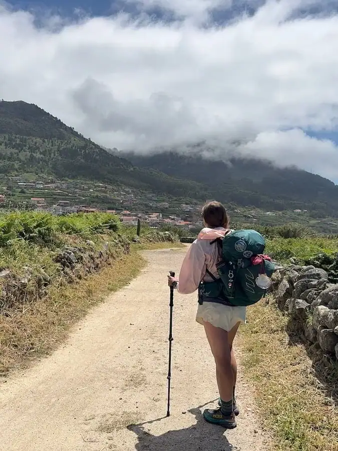 Mikala standing on a dirt path with her backpack and hiking poles looking away from the camera back towards a mountain and town in the distance while hiking solo on the Camino Portugués Coastal route.
