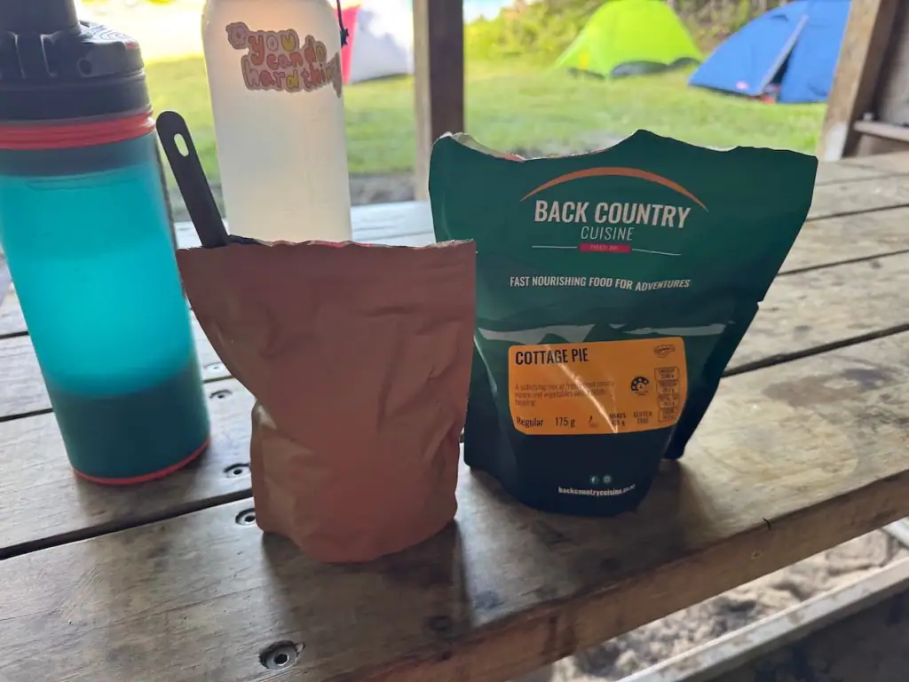 Freeze dried hiking meal, water bottles, and a cooking pouch set up on a wooden campsite table with tents visible outside in the background on the Rakiura Track.