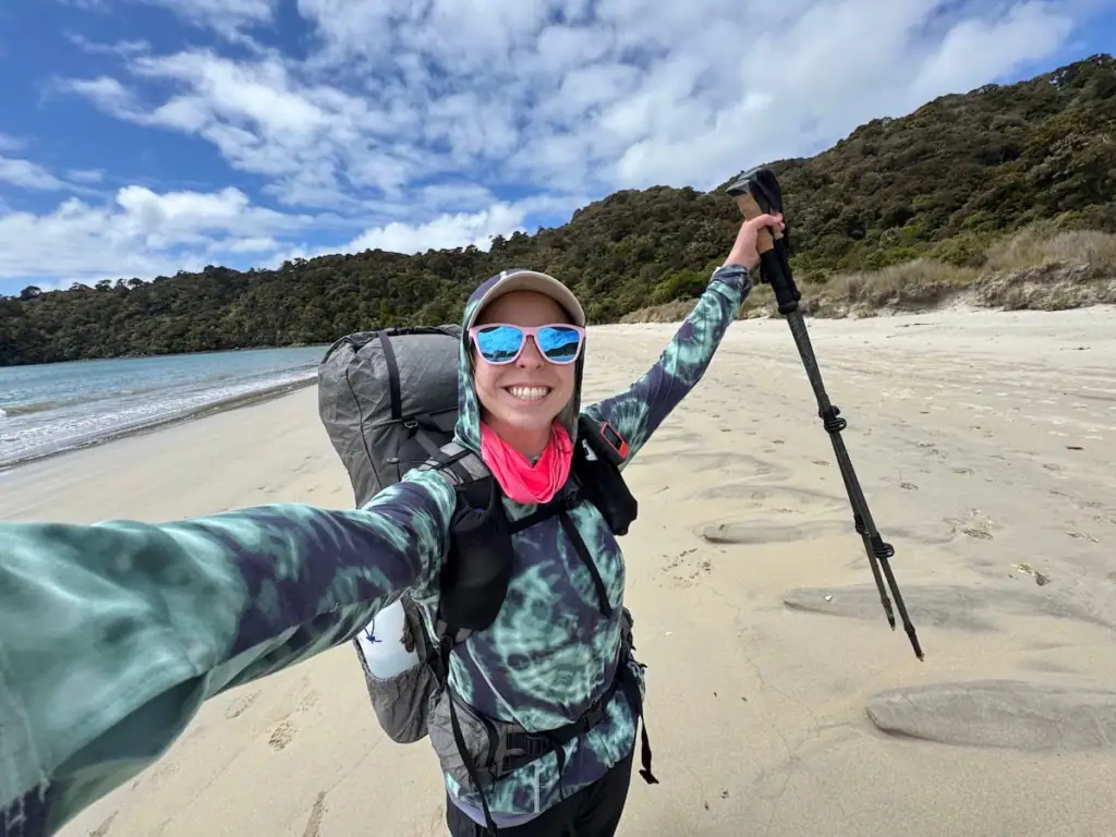 Mikala taking a selfie on the Rakiura Track while hiking along a sandy beach with a large backpack and trekking pole raised in celebration. Her layered sun protection and backpack setup naturally show essentials for an NZ Great Walks packing list.