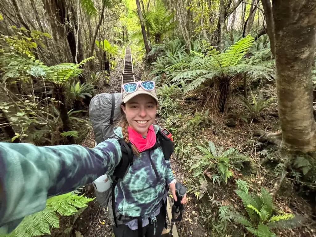 Mikala smiles at the camera while taking a selfie on the Rakiura Track, wearing a backpacking pack, hiking gear, and sunglasses perched on her cap.