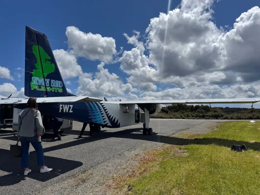 A passenger walks toward a small Stewart Island Flights aircraft parked on the tarmac beneath towering white clouds and a bright blue sky. Flying to Stewart Island offers a scenic and efficient alternative to the ferry, with the adventure beginning before takeoff.