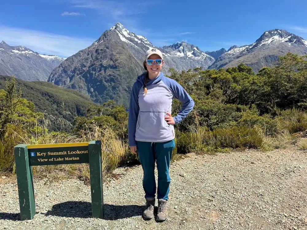 Hiker standing beside a sign reading "Key Summit Lookout. View of Lake Marian" with snow capped mountains in the background near the Routeburn Track. Bright sunshine and clear skies highlight the alpine viewpoint.