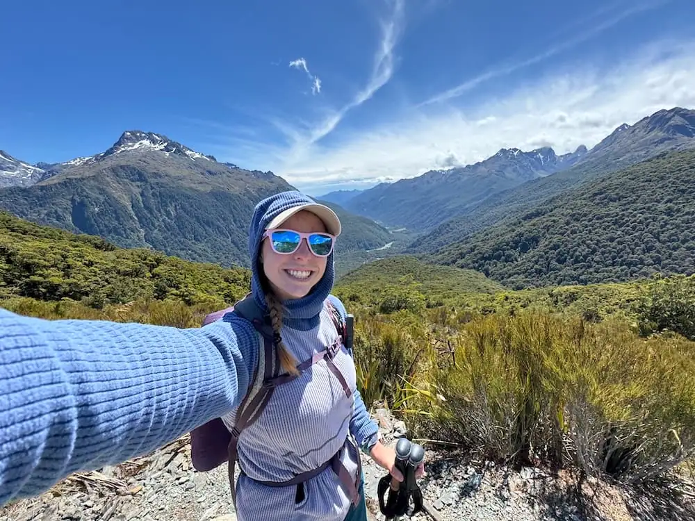 Smiling hiker wearing a backpack pauses for a selfie overlooking a wide mountain valley while hiking to Key Summit. Snow capped peaks and dense forest stretch into the distance beneath wispy clouds.