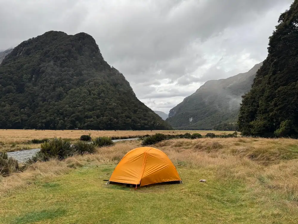 Bright orange tent pitched in an open grassy valley surrounded by forested mountains and dark cloudy skies at the Routeburn Flats Campsite on the Routeburn Track. A small stream winds through the landscape, adding to the remote backcountry setting.