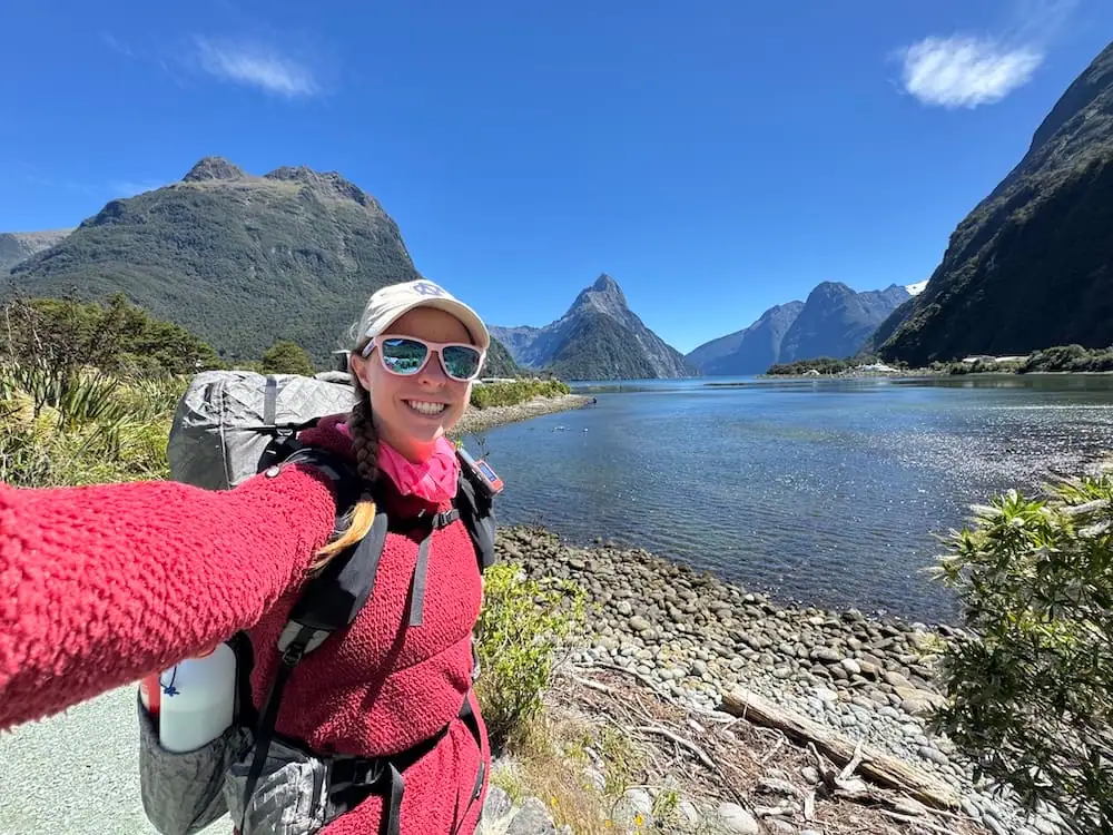Mikala taking a selfie at Milford Sound beside calm water surrounded by steep mountains on a sunny day while wearing hiking gear and carrying a backpack. Her layered clothing, sunglasses, and trail setup naturally reflect useful gear for an NZ Great Walks packing list.