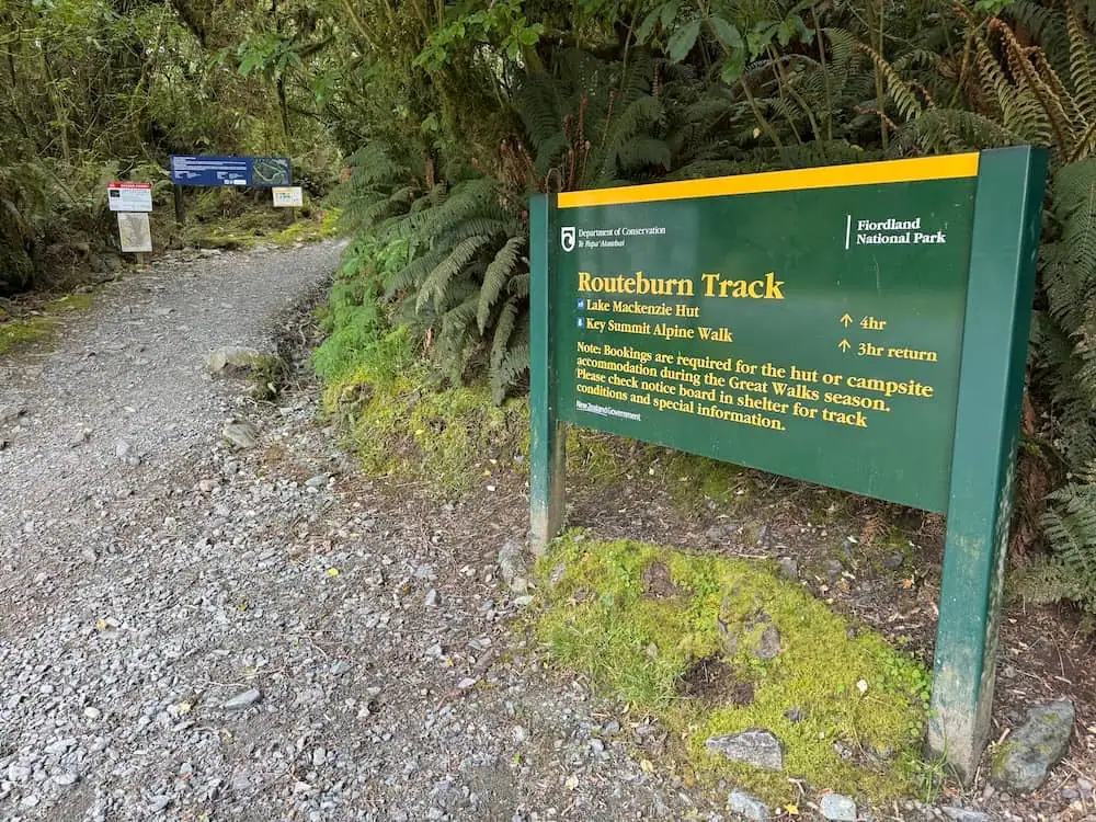 Trailhead sign for the Routeburn Track standing beside a rocky forest path surrounded by moss and ferns. The sign reads "Routeburn Track" with directions to Lake Mackenzie Hut and Key Summit Alpine Walk. This sign marks the start of the track on The Divide Shelter side, but the track can be walked in either direction.