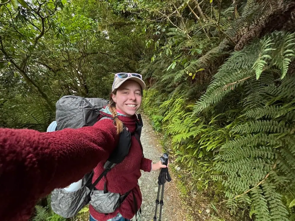Smiling hiker wearing a backpack and holding trekking poles walks through lush green forest on the Routeburn Track. Ferns line the narrow trail, showing the dense rainforest scenery of the lower sections.