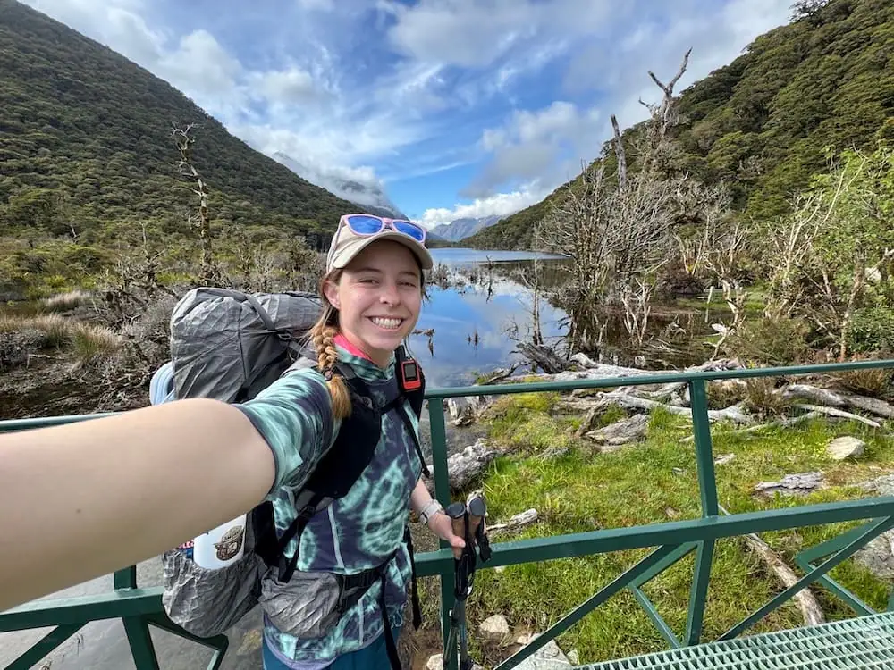 Smiling hiker with a backpack takes a selfie on a bridge overlooking a calm Lake Howden surrounded by forested mountains on the Routeburn Track. Reflections shimmer across the water as clouds gather above the valley.